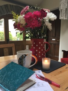 A jug of flowers, a lit candle, a mug of coffee, a notebook and a pen on a kitchen table