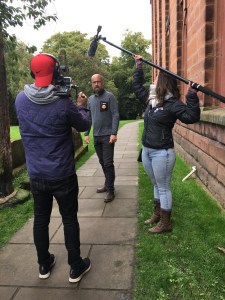 a man with a camera filming another man talking, while a woman holds a boom microphone over the scene